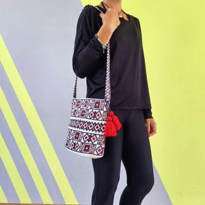 May include: A white, red, and navy blue patterned crossbody bag with red tassels. The bag has a woven design and a matching strap. The bag is held by a person wearing a black long-sleeve shirt and black leggings.