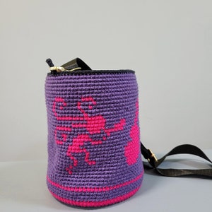 May include: Purple crocheted bucket bag with a pink floral design and a black adjustable strap.