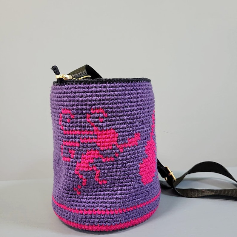 May include: Purple crocheted bucket bag with a pink floral design and a black adjustable strap.