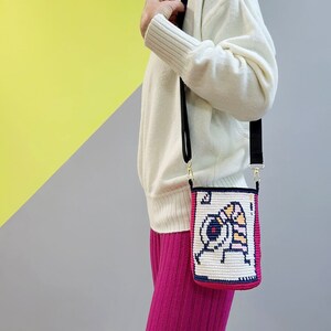 May include: A white crocheted crossbody bag with a black strap and a colorful pixelated design. The bag is worn over a pink sweater and pink pants.