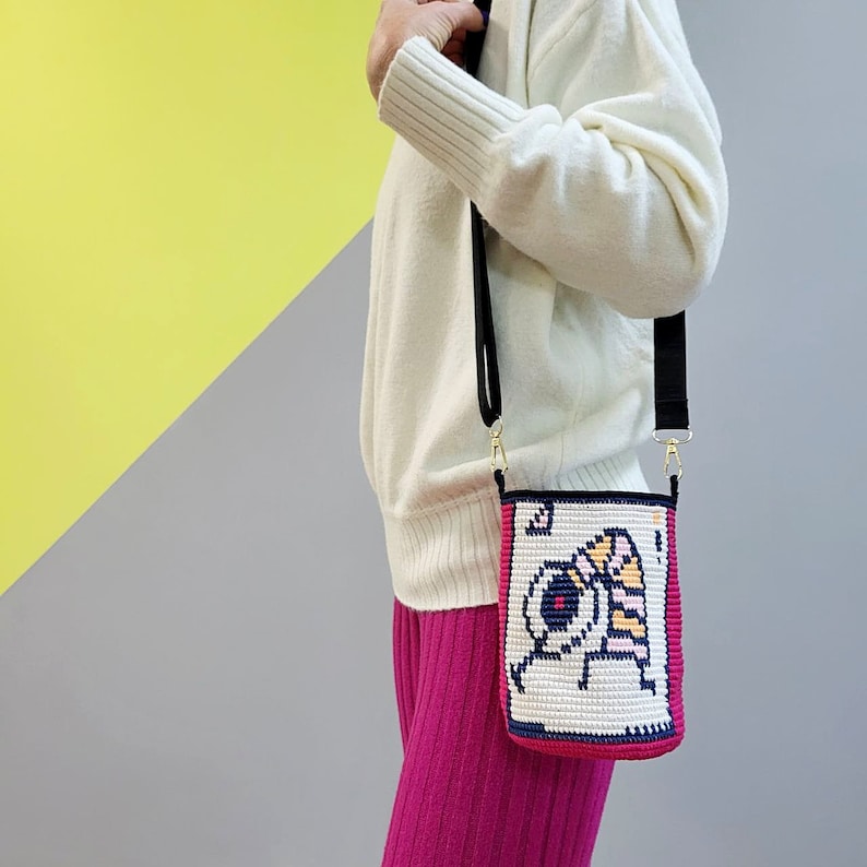May include: A white crocheted crossbody bag with a black strap and a colorful pixelated design. The bag is worn over a pink sweater and pink pants.