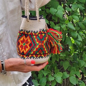 May include: A small, cylindrical, crocheted bucket bag featuring a cream-colored base with a vibrant geometric pattern in red, yellow, and green. The bag includes two braided handles and a large, multi-colored yarn tassel attached to the side.
