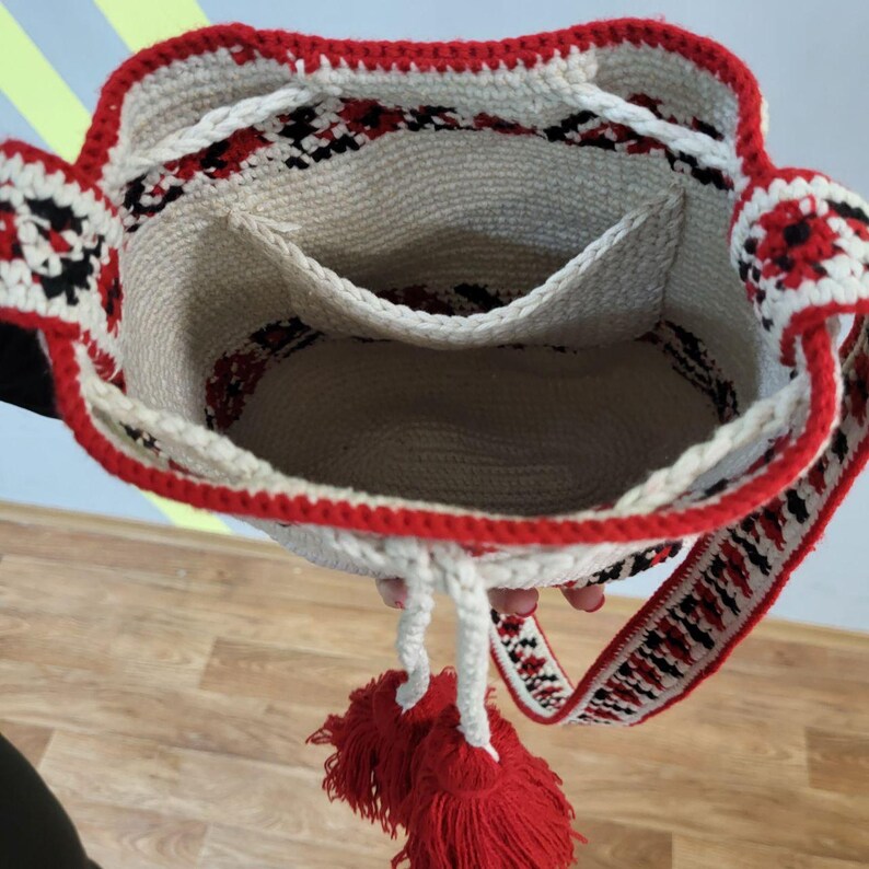 May include: A white crocheted bag with red and black trim. The bag has a drawstring closure and a long strap with tassels. The bag has a small pocket on the inside.