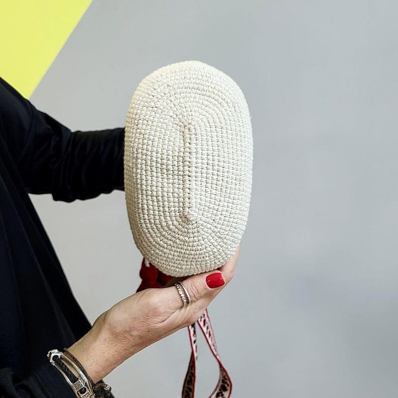 May include: A white crocheted bag with a red and white patterned strap. The bag is oval shaped and has a textured surface.