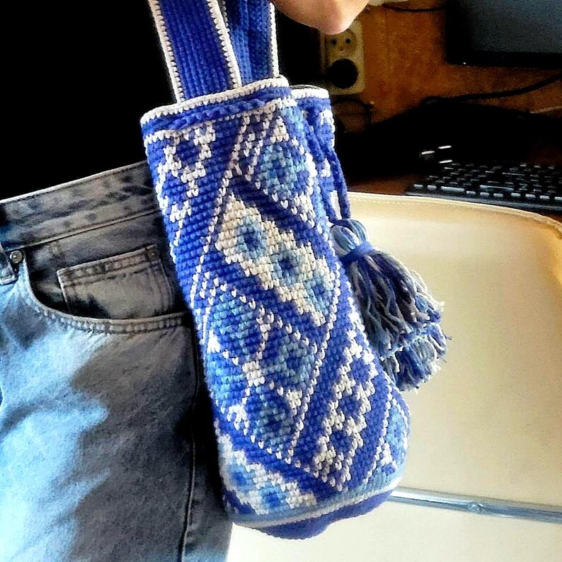 A blue and white crocheted bag with a geometric pattern. The bag has a long strap and a tassel. The bag is held by a person wearing blue jeans.