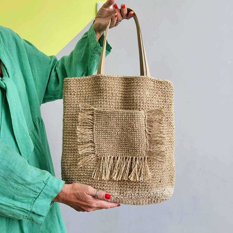 Front view of natural jute crochet shoulder tote showing the large exterior pocket with decorative fringe.