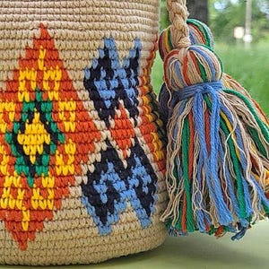 May include: A beige crochet bucket bag featuring a geometric pattern in orange, yellow, green, and blue. The bag includes a thick braided strap and a large, multi-colored tassel made of blue, green, orange, yellow, and beige yarn strands.