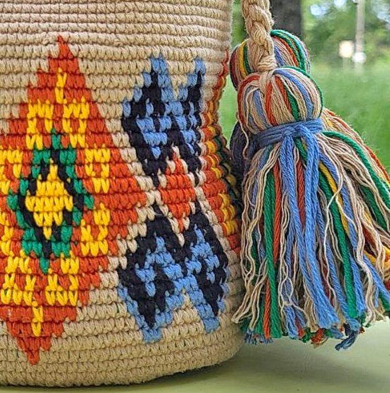 May include: A beige crochet bucket bag featuring a geometric pattern in orange, yellow, green, and blue. The bag includes a thick braided strap and a large, multi-colored tassel made of blue, green, orange, yellow, and beige yarn strands.