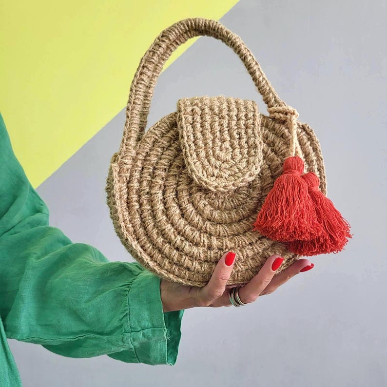 May include: A round, beige, crocheted handbag made of natural fiber, featuring a circular pattern, a top handle, a flap closure, and a pair of bright red decorative tassels attached to the side. The bag has a textured, woven appearance.