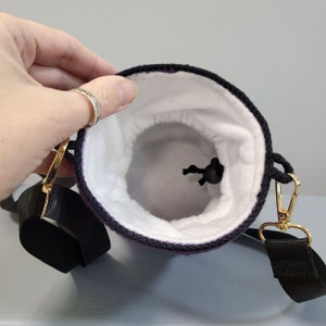 May include: Purple crocheted drawstring bag with a black strap and a white fleece lining. The bag has a gold metal clasp and a black drawstring.