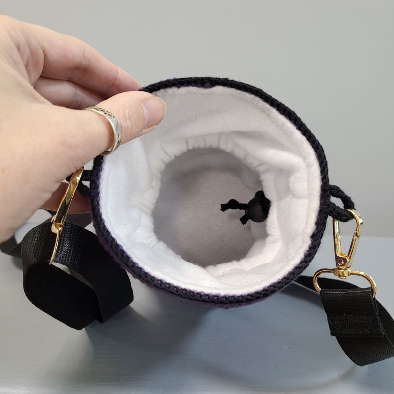 May include: Purple crocheted drawstring bag with a black strap and a white fleece lining. The bag has a gold metal clasp and a black drawstring.