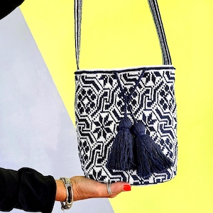 May include: A navy blue and white patterned bucket bag with a matching strap and two navy blue tassels. The bag is held up against a yellow and gray background. The bag's pattern is geometric.