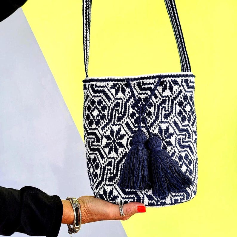 May include: A navy blue and white patterned bucket bag with a matching strap and two navy blue tassels. The bag is held up against a yellow and gray background. The bag's pattern is geometric.