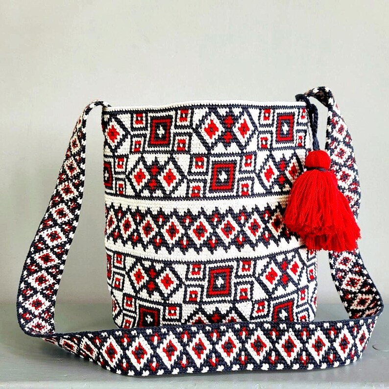 May include: A white, red, and navy blue patterned crossbody bag with a matching strap. The bag features a geometric design and a red tassel. The strap is adjustable. The bag is made of woven material.