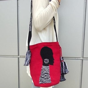 May include: Red crocheted tote bag with a black silhouette of a person's head with a white and black striped pattern. The bag has black and white tassels.