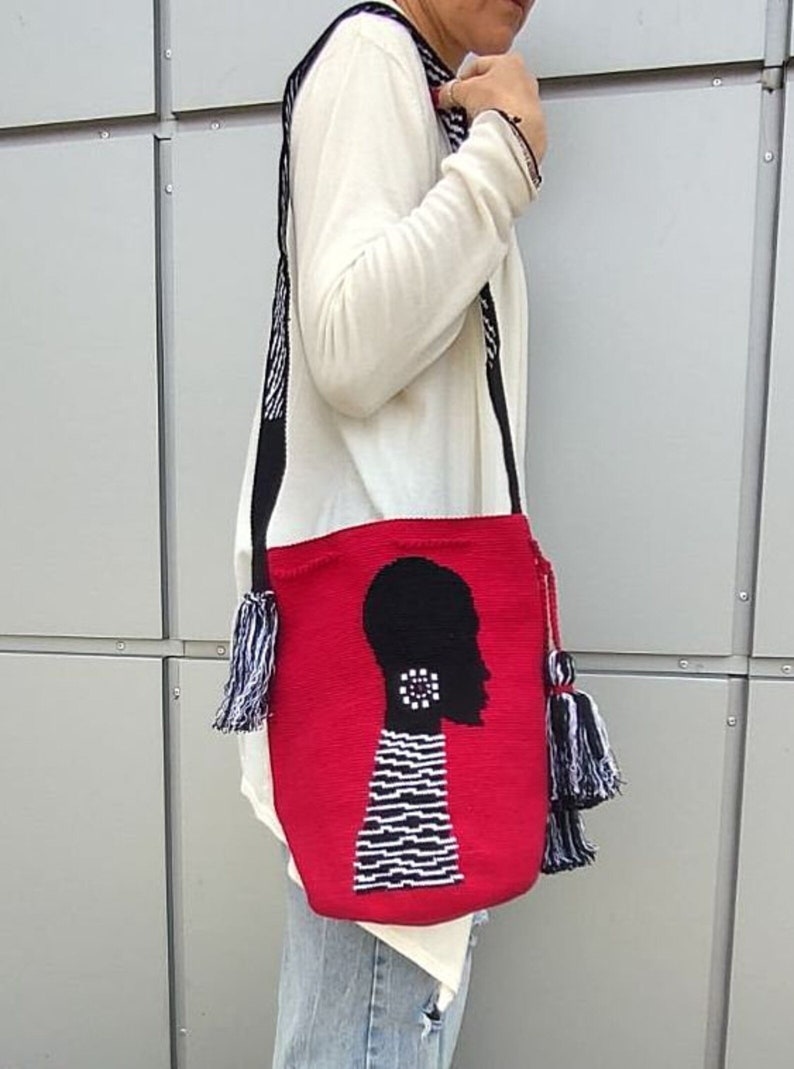 May include: Red crocheted tote bag with a black silhouette of a person's head with a white and black striped pattern. The bag has black and white tassels.