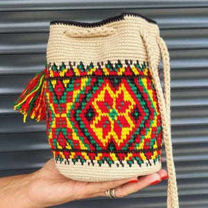 May include: A beige crochet bucket bag featuring a vibrant geometric pattern in red, yellow, and green. The bag includes a braided drawstring closure, a matching braided shoulder strap, and a decorative tassel with red, yellow, and green yarn.