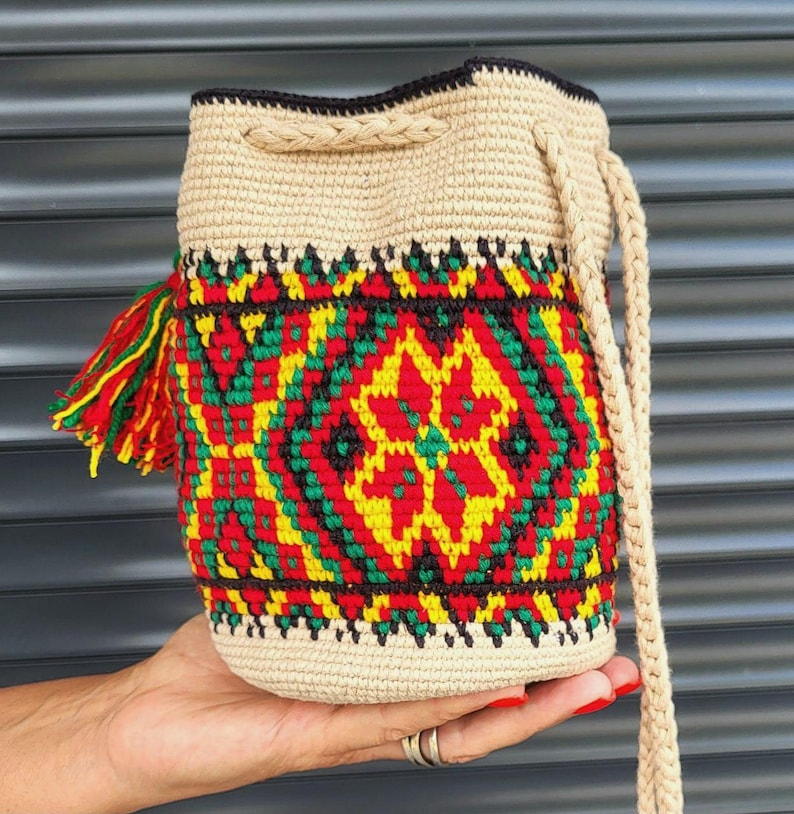 May include: A beige crochet bucket bag featuring a vibrant geometric pattern in red, yellow, and green. The bag includes a braided drawstring closure, a matching braided shoulder strap, and a decorative tassel with red, yellow, and green yarn.