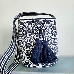 May include: A navy blue and white woven shoulder bag with a geometric pattern. The bag has a long strap with a striped design and two navy blue tassels. The bag is cylindrical in shape.