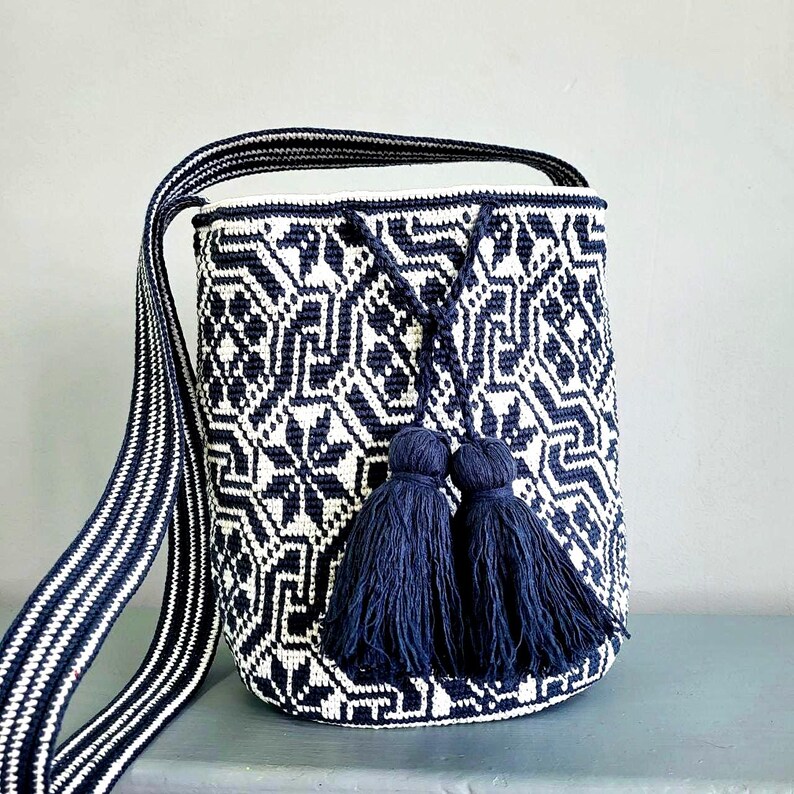 May include: A navy blue and white woven shoulder bag with a geometric pattern. The bag has a long strap with a striped design and two navy blue tassels. The bag is cylindrical in shape.