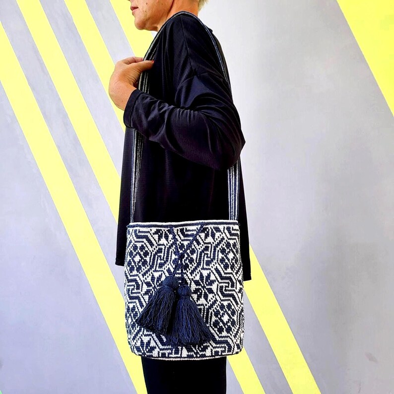 May include: A black and white geometric patterned shoulder bag with two navy blue tassels. The bag has a long strap and is worn over the shoulder. The bag is made of woven material.