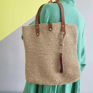 May include: A rectangular tote bag crafted from tan woven material, featuring two brown leather shoulder straps secured with silver-toned metal rivets. A central snap closure secures the top, and a brown leather tassel hangs from one strap.