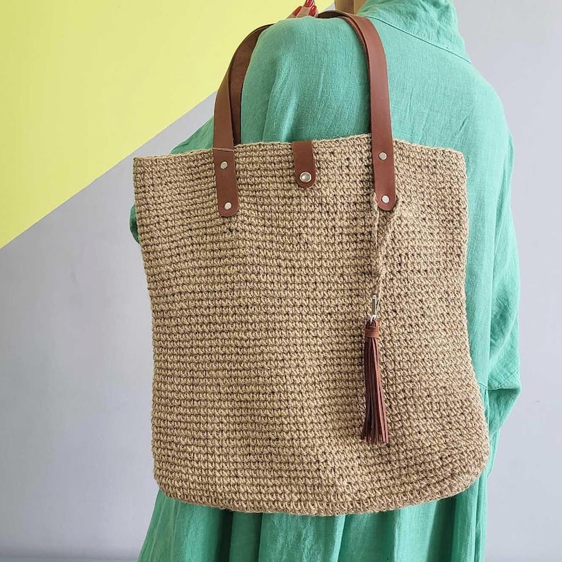 May include: A rectangular tote bag crafted from tan woven material, featuring two brown leather shoulder straps secured with silver-toned metal rivets. A central snap closure secures the top, and a brown leather tassel hangs from one strap.