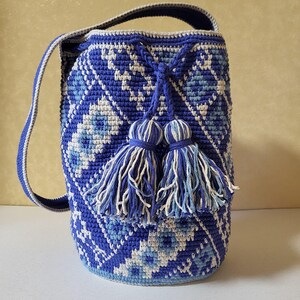 A handmade blue crochet bucket bag with a geometric pattern in white and light blue. The bag has a drawstring closure with two matching tassels and a shoulder strap. The bag is approximately 10 inches tall.