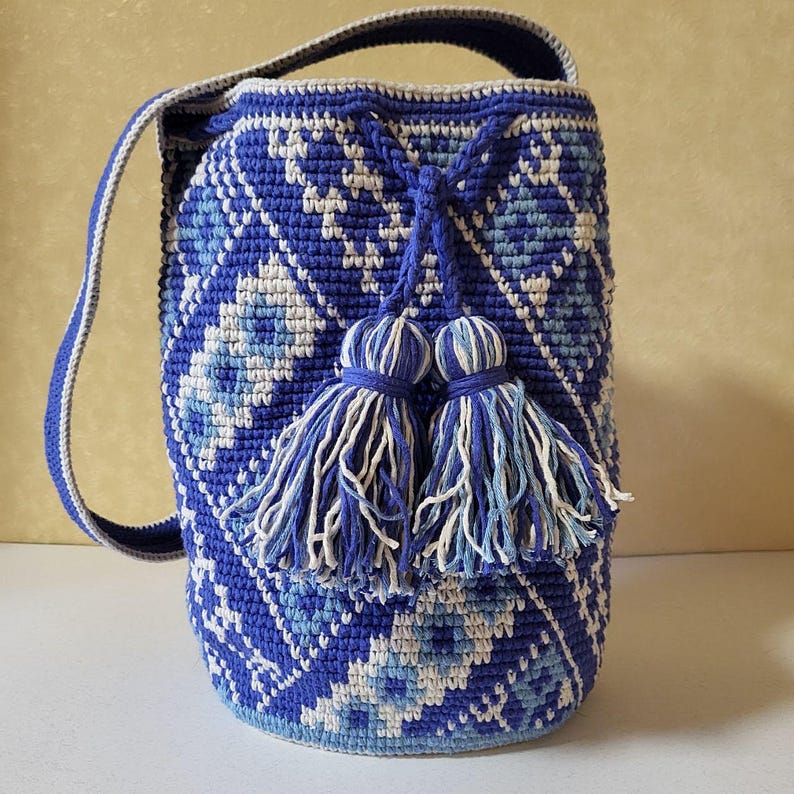 A handmade blue crochet bucket bag with a geometric pattern in white and light blue. The bag has a drawstring closure with two matching tassels and a shoulder strap. The bag is approximately 10 inches tall.