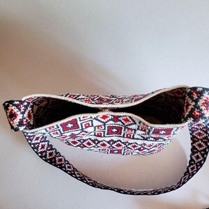 May include: A handmade shoulder bag with a geometric pattern in red, white, and navy blue. The bag features a zippered top and a long strap. The interior is visible, showing a dark fabric lining. The bag's design is a repeating pattern of squares and diamonds.