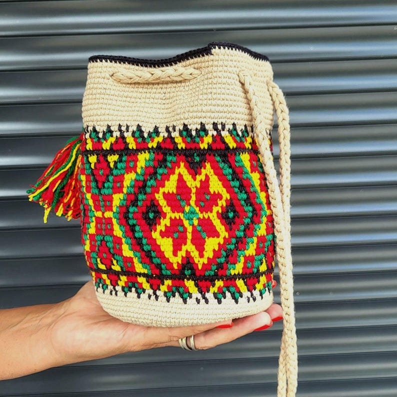 May include: A beige crochet bucket bag featuring a vibrant geometric pattern in red, yellow, and green. The bag includes a drawstring closure with a braided cord and a matching tassel on the side, showcasing intricate needlework and texture.