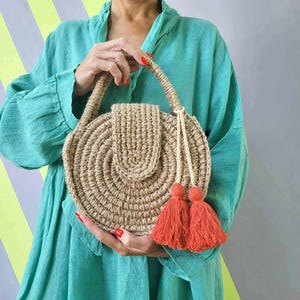 May include: A round, crocheted jute handbag featuring a structured top handle and a flap closure. The bag is decorated with two vibrant orange-red tassels hanging from the handle, adding a pop of color to the natural tan, textured fiber construction.