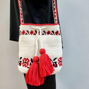 May include: White crocheted tote bag with red and black floral embroidery and red tassels. The bag has a long strap.