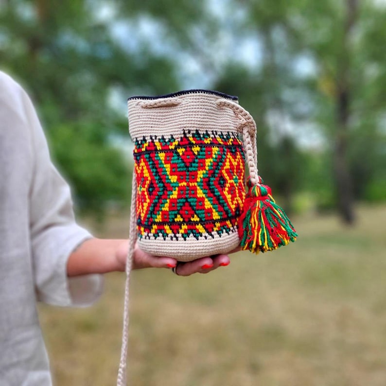May include: A cylindrical crochet bucket bag in a light beige tone, featuring a vibrant geometric pattern in red, yellow, and green. The bag includes a matching braided shoulder strap and a decorative tassel in the same color palette attached.