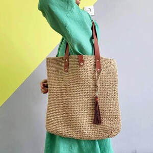 May include: A large, rectangular tote bag crafted from a natural tan woven fiber material. The bag features two brown leather shoulder straps secured with silver-toned metal rivets and a matching brown leather tassel hanging from one of the straps.