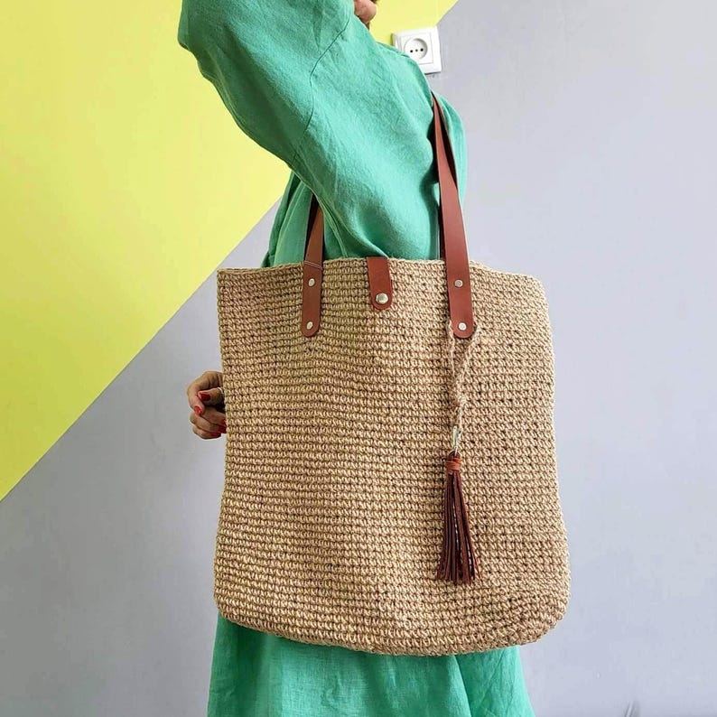 May include: A large, rectangular tote bag crafted from a natural tan woven fiber material. The bag features two brown leather shoulder straps secured with silver-toned metal rivets and a matching brown leather tassel hanging from one of the straps.