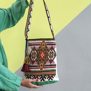 May include: A white woven shoulder bag featuring a vibrant, multi-colored geometric diamond pattern in shades of orange, green, black, and red. The bag includes a long, matching patterned strap and a decorative tassel in yellow and dark red.