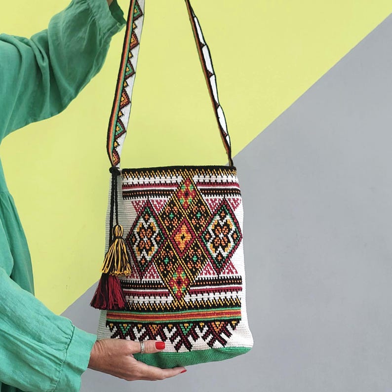 May include: A white woven shoulder bag featuring a vibrant, multi-colored geometric diamond pattern in shades of orange, green, black, and red. The bag includes a long, matching patterned strap and a decorative tassel in yellow and dark red.