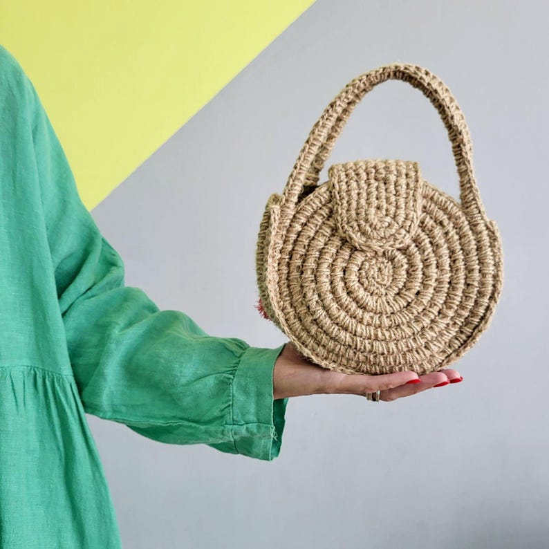 May include: A round, tan, crocheted handbag made of natural fiber, featuring a circular spiral pattern, a structured top handle, and a small flap closure. The bag has a textured, woven appearance and is held in a palm, showcasing its compact size.