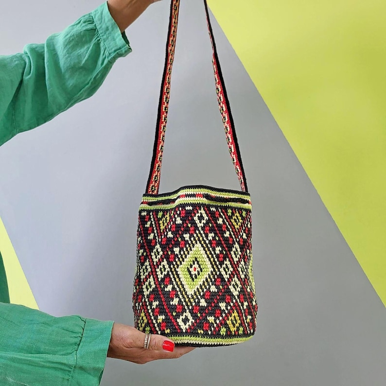 May include: A cylindrical crochet bucket bag featuring a black base with a geometric diamond pattern in lime green, red, and white. The bag includes a long, matching patterned shoulder strap and a reinforced black rim at the top opening.
