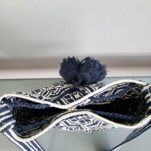 May include: A navy blue and white woven bag with a geometric pattern. The bag has a striped strap and a navy blue tassel. The bag is open, revealing the interior.