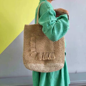May include: A tan woven straw tote bag featuring a square front pocket with frayed fringe detailing along the edges. The bag has two light-colored shoulder straps and a textured, natural fiber construction with a rectangular silhouette.