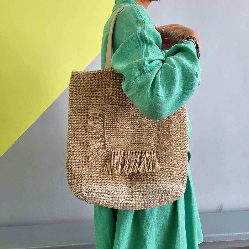 May include: A tan woven straw tote bag featuring a square front pocket with frayed fringe detailing along the edges. The bag has two light-colored shoulder straps and a textured, natural fiber construction with a rectangular silhouette.