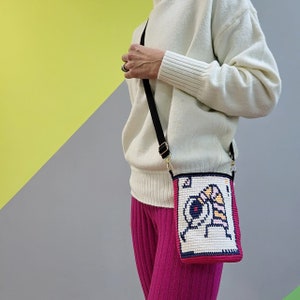 May include: A white sweater with a black and gold crossbody bag. The bag has a white and pink embroidered design with a cartoon character.