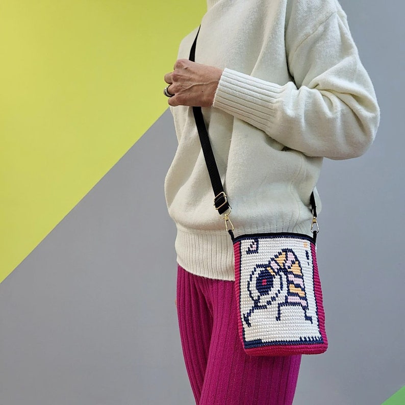 May include: A white sweater with a black and gold crossbody bag. The bag has a white and pink embroidered design with a cartoon character.