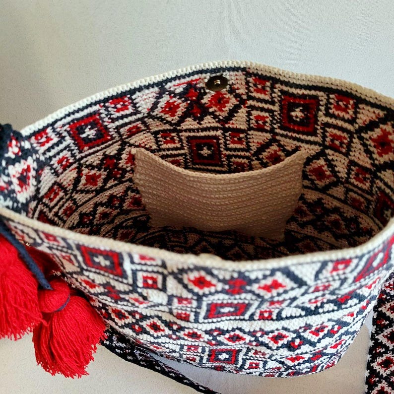 May include: A handmade crochet bag with a geometric pattern in red, black, and white. The bag features a cream-colored interior pocket and is adorned with red tassels. The strap is visible on the right side of the bag.