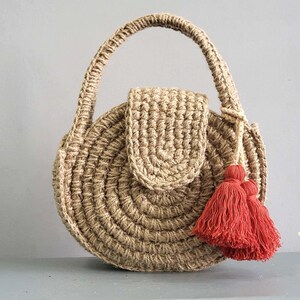 May include: A round, tan, crocheted jute handbag featuring a structured top handle and a central flap closure. The bag is accented with a pair of vibrant, deep red decorative tassels attached to the side, creating a distinct contrast against the natural, textured fiber of the circular body.