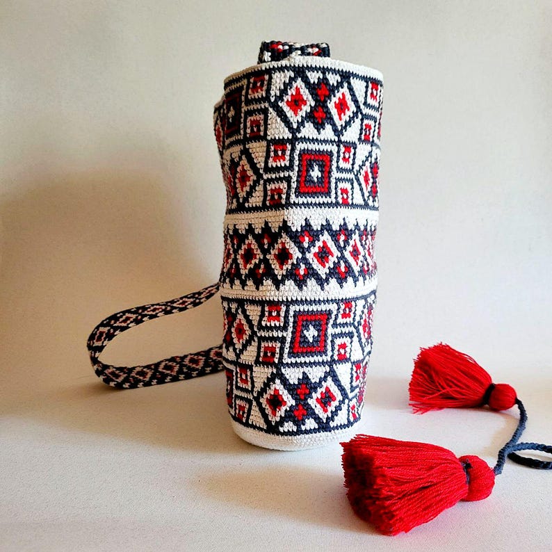 May include: A cylindrical white, red, and navy blue crocheted bag with a geometric pattern. The bag has a matching strap and two red tassels. The bag is approximately 12 inches tall.
