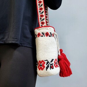 May include: White crocheted drawstring bag with red and black floral embroidery. The bag has a long red, white and black striped strap with tassels.