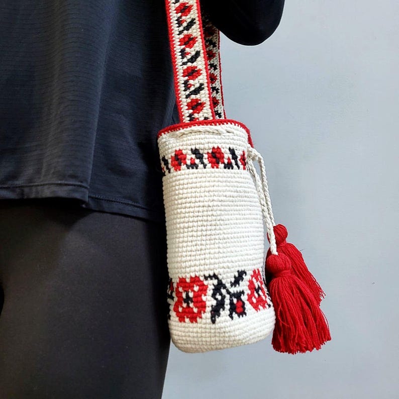 May include: White crocheted drawstring bag with red and black floral embroidery. The bag has a long red, white and black striped strap with tassels.